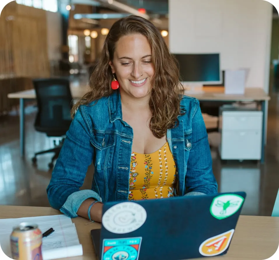 Woman in yellow shirt and jean jacket smiling at her laptop