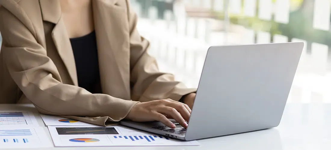 Woman in blazer typing on laptop with papers showing charts and graphs on desk